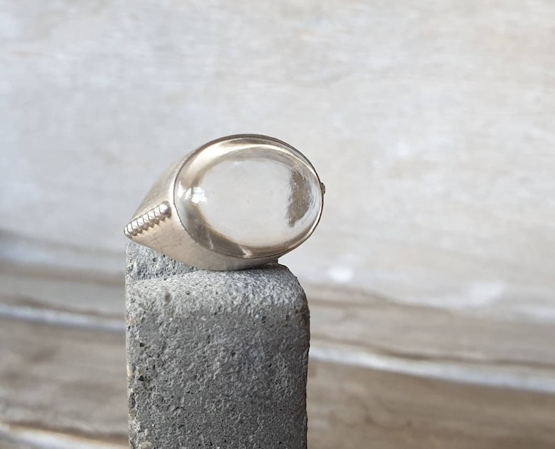 May include: A silver signet ring with a clear, oval-shaped stone. The ring is set on a gray, textured surface. The ring's band has a decorative, ridged design on the side.