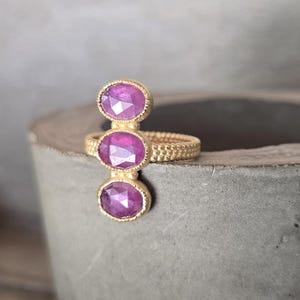 May include: A gold ring featuring three vertically aligned, oval-shaped, faceted magenta gemstones. The band of the ring has a textured, beaded surface, and each of the three stones is set within a delicate, beaded gold bezel setting.