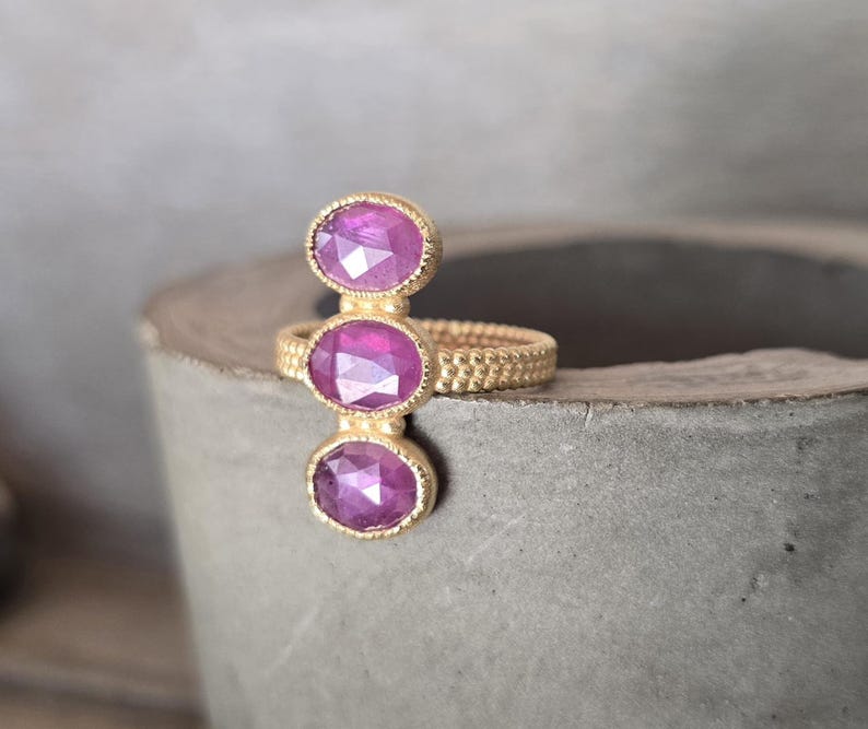 May include: A gold ring featuring three vertically aligned, oval-shaped, faceted magenta gemstones. The band of the ring has a textured, beaded surface, and each of the three stones is set within a delicate, beaded gold bezel setting.