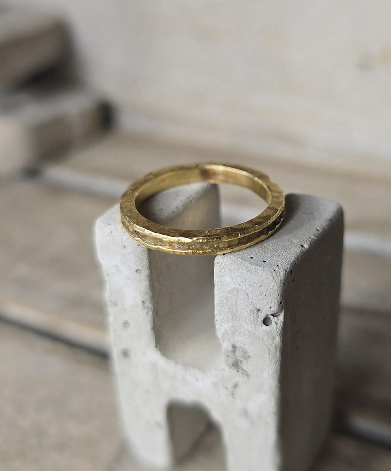 Pu&ograve; includere: Un anello a fascia d'oro con una texture martellata. L'anello &egrave; appoggiato su un blocco di cemento.