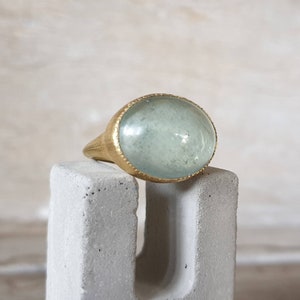 May include: A gold ring with a large oval aquamarine gemstone. The ring is set on a gray concrete surface.