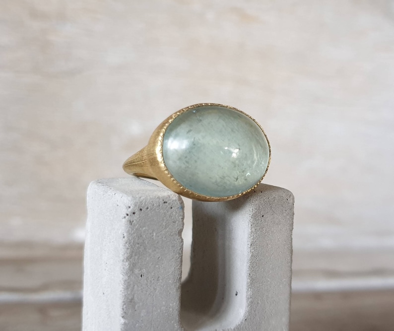 May include: A gold ring with a large oval aquamarine gemstone. The ring is set on a gray concrete surface.