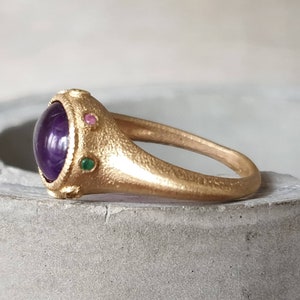 K&ouml;nnte beinhalten: Ein goldener Ring mit einem gro&szlig;en violetten Edelstein, umgeben von kleineren gr&uuml;nen und rosa Edelsteinen.