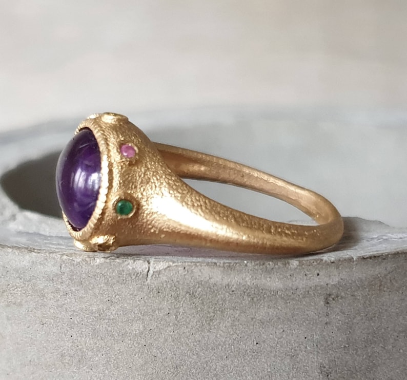 K&ouml;nnte beinhalten: Ein goldener Ring mit einem gro&szlig;en violetten Edelstein, umgeben von kleineren gr&uuml;nen und rosa Edelsteinen.