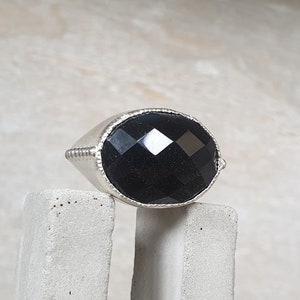 May include: A silver ring with a large, faceted black gemstone. The ring is sitting on a gray concrete surface.