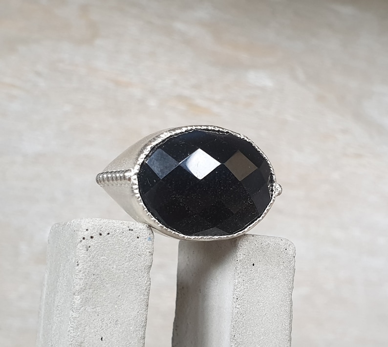 May include: A silver ring with a large, faceted black gemstone. The ring is sitting on a gray concrete surface.
