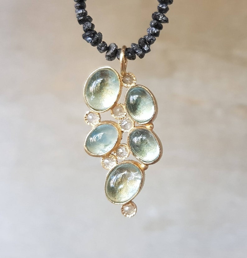 Pu&ograve; includere: Una delicata collana d'oro con un ciondolo che presenta un gruppo di pietre di acquamarina blu chiaro e piccoli diamanti trasparenti. La collana &egrave; realizzata con perline nere.