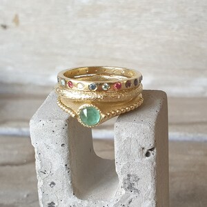 May include: A collection of gold rings with gemstone accents. The rings are stacked on a gray concrete block. One ring features a large, round, green gemstone. Other rings have small, colorful gemstones. The rings have a textured, detailed design.