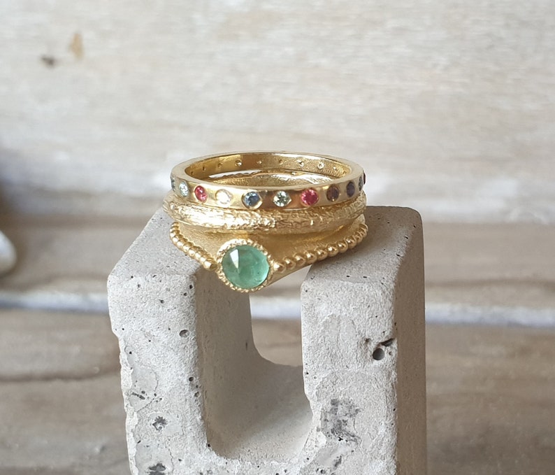 May include: A collection of gold rings with gemstone accents. The rings are stacked on a gray concrete block. One ring features a large, round, green gemstone. Other rings have small, colorful gemstones. The rings have a textured, detailed design.