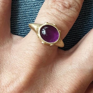 K&ouml;nnte beinhalten: Ein goldener Ring mit einem violetten Edelstein, der in einer Zargenfassung gefasst ist. Der Ring hat eine strukturierte Band und ein dekoratives Design um den Edelstein.
