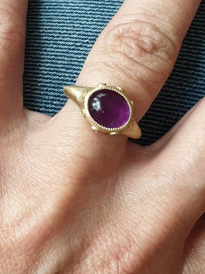 K&ouml;nnte beinhalten: Ein goldener Ring mit einem violetten Edelstein, der in einer Zargenfassung gefasst ist. Der Ring hat eine strukturierte Band und ein dekoratives Design um den Edelstein.