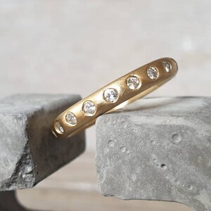 Peut inclure: Une bague en or avec une finition mate, sertie de plusieurs petites pierres pr&eacute;cieuses claires. La bague repose sur une surface grise et textur&eacute;e, mettant en valeur son design. L'anneau de la bague est &eacute;pais et arrondi.