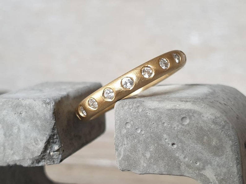 Peut inclure: Une bague en or avec une finition mate, sertie de plusieurs petites pierres pr&eacute;cieuses claires. La bague repose sur une surface grise et textur&eacute;e, mettant en valeur son design. L'anneau de la bague est &eacute;pais et arrondi.