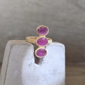 May include: A gold ring featuring a textured, beaded band and three vertically aligned, oval-shaped, faceted pink gemstones. The stones are set in gold bezels, creating a linear design that extends upwards from the centre of the ring band.