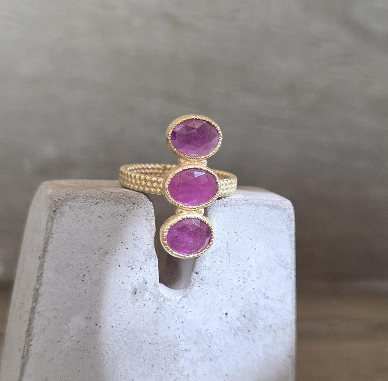 May include: A gold ring featuring a textured, beaded band and three vertically aligned, oval-shaped, faceted pink gemstones. The stones are set in gold bezels, creating a linear design that extends upwards from the centre of the ring band.