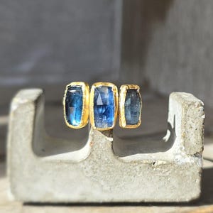May include: Three blue gemstone rings set in gold. The rings are arranged in a row, with the middle ring slightly larger than the other two.