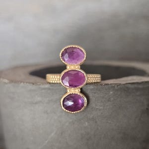 May include: A gold ring featuring three vertically aligned, oval-cut purple gemstones. The band has a textured, beaded surface, and each gemstone is set in a delicate, scalloped gold bezel setting that highlights the faceted surface of the stones.