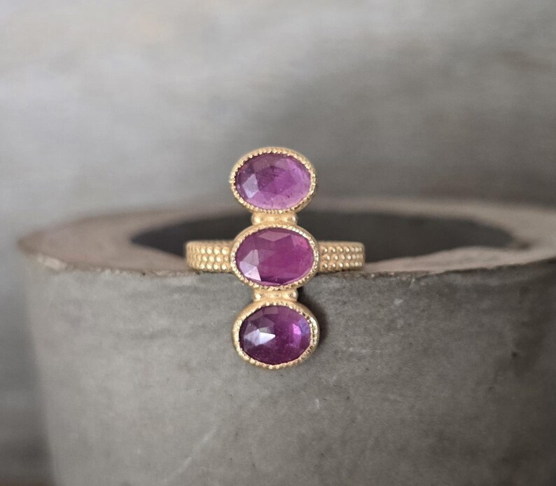 May include: A gold ring featuring three vertically aligned, oval-cut purple gemstones. The band has a textured, beaded surface, and each gemstone is set in a delicate, scalloped gold bezel setting that highlights the faceted surface of the stones.