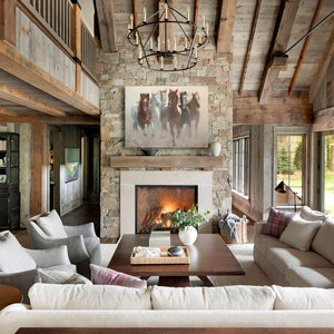 May include: A large living room with a stone fireplace and a painting of horses above it. The room has a vaulted ceiling with exposed wooden beams and large windows. There are several couches and chairs arranged around a coffee table.