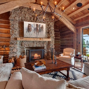 May include: A rustic living room with a stone fireplace, a large wooden coffee table, and a white couch. The room has a large window with a view of a lake. A painting of horses hangs above the fireplace.