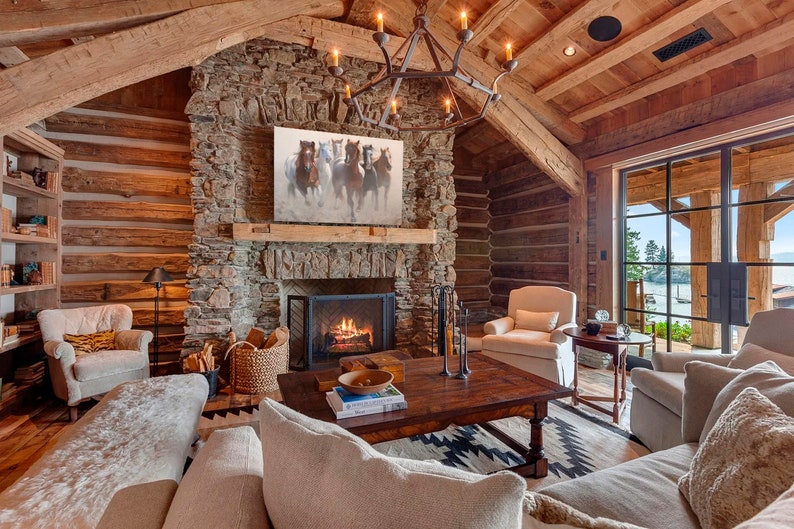 May include: A rustic living room with a stone fireplace, a large wooden coffee table, and a white couch. The room has a large window with a view of a lake. A painting of horses hangs above the fireplace.