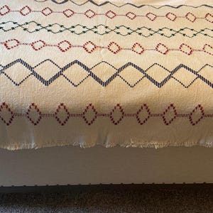 May include: Cream-colored throw blanket with embroidered geometric patterns in red, green, and navy blue. The blanket has a fringed edge and is draped over a white upholstered bench. The patterns include diamond and zigzag designs.