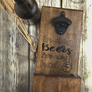 May include: Wooden bottle opener with a black bottle opener attached. The bottle opener has the text "In dog beers I've only had one" with a black hand pointing up with one finger.
