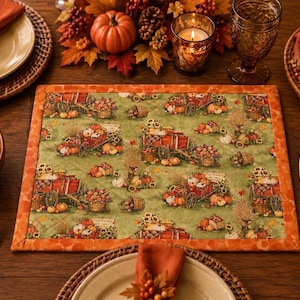 May include: A rectangular quilted placemat featuring a green fabric background with a repeating pattern of red wagons, pumpkins, sunflowers, and baskets. The placemat is finished with a wide orange border featuring a subtle tonal print.