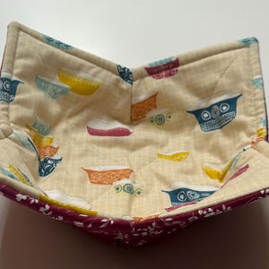 May include: A quilted fabric bowl with a burgundy exterior and a cream interior. The interior features a pattern of colorful bowls.