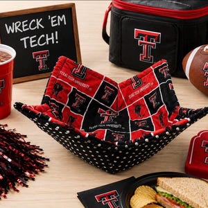 May include: A fabric bowl cozy featuring a red and black Texas Tech University logo pattern, paired with a red plastic cup, a black insulated lunch bag, a football, a foam finger, a chalkboard sign reading WRECK 'EM TECH!, and black pom-poms.
