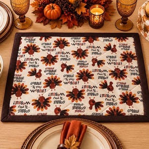 May include: A rectangular quilted placemat featuring a repeating pattern of brown turkeys and the text "GOBBLE 'TIL YOU WOBBLE!" on an off-white background, finished with a solid dark brown fabric border along all four edges of the quilted fabric.