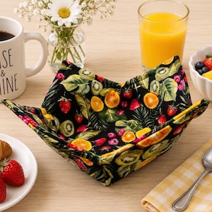 May include: A square fabric bowl cozy featuring a black background with a vibrant, colorful print of various fruits including strawberries, kiwi slices, and orange segments. The quilted holder has four folded corners and is designed to hold a bowl.