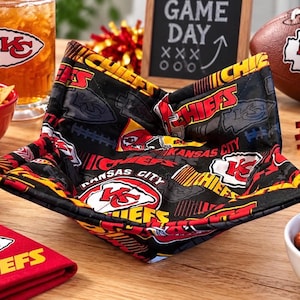 May include: A fabric bowl cozy featuring a black background with a repeating Kansas City Chiefs logo, red and yellow text, and football-themed graphics. The square-shaped holder has folded corners and stitched seams, designed to hold a bowl securely.