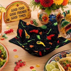 May include: A black fabric bowl cozy featuring a colorful pattern of red, yellow, and green chili peppers. The square-shaped, quilted holder has four folded corners and is designed to hold a bowl, showcasing a vibrant pepper print throughout.