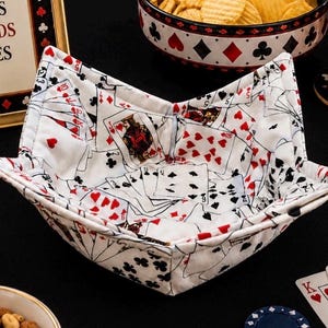 May include: A fabric bowl cozy featuring a white background with a repeating pattern of playing cards, including hearts, diamonds, clubs, and spades. The square-shaped bowl holder has four folded corners and stitched seams, designed to hold a bowl securely.
