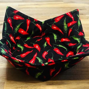 May include: A black fabric bowl with a red and green chili pepper pattern. The bowl is square-shaped with folded corners, designed to hold food. The peppers are scattered across the surface.