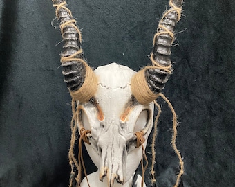 May include: A white skull mask with black and silver horns and brown tassels. The mask has a tribal design and is perfect for a costume or cosplay.