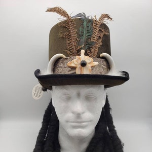 Puede incluir: Un sombrero alto de copa verde con una banda negra y un adorno de hueso. El sombrero está decorado con plumas y tiene una piedra oscura en el centro del adorno.