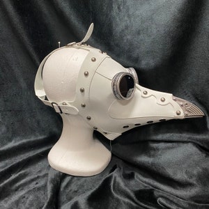 Peut inclure: Masque de m&eacute;decin de la peste en cuir blanc avec des accents argent&eacute;s et deux pi&egrave;ces rondes pour les yeux. Le masque est sur une t&ecirc;te de mannequin blanche.