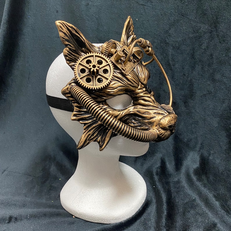 May include: A gold and brown steampunk-style fox mask with intricate gears and a pipe-like detail. The mask is mounted on a white mannequin head.