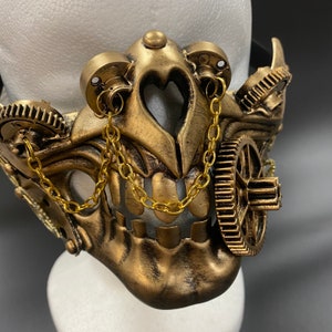Peut inclure: Un masque de style steampunk de couleur dor&eacute;e avec des d&eacute;tails complexes, notamment des engrenages, des cha&icirc;nes et un motif de cr&acirc;ne.