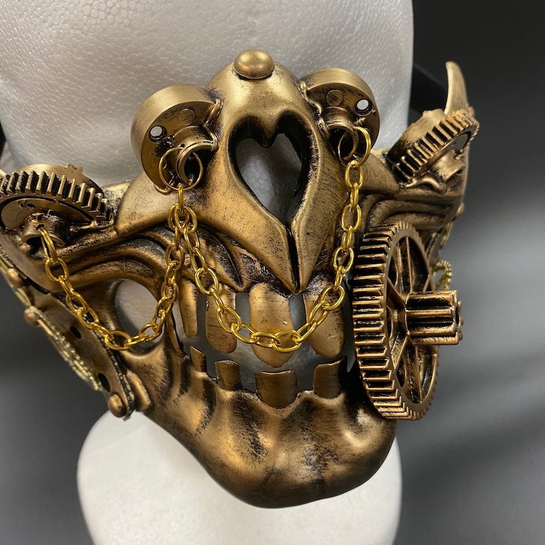Peut inclure: Un masque de style steampunk de couleur dor&eacute;e avec des d&eacute;tails complexes, notamment des engrenages, des cha&icirc;nes et un motif de cr&acirc;ne.