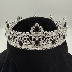 May include: Silver-toned crown with intricate filigree and black gemstone accents. The crown features a detailed design with floral and leaf patterns, and is set against a black background. A regal accessory.