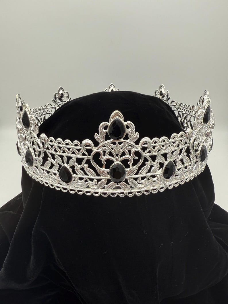 May include: Silver-toned crown with intricate filigree and black gemstone accents. The crown features a detailed design with floral and leaf patterns, and is set against a black background. A regal accessory.