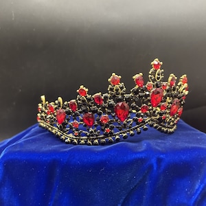 May include: A gold crown with red and black rhinestones. The crown is on a blue velvet surface.