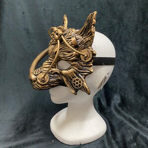 May include: A gold and black steampunk-style mask with gears and pipes. The mask is on a white mannequin head.