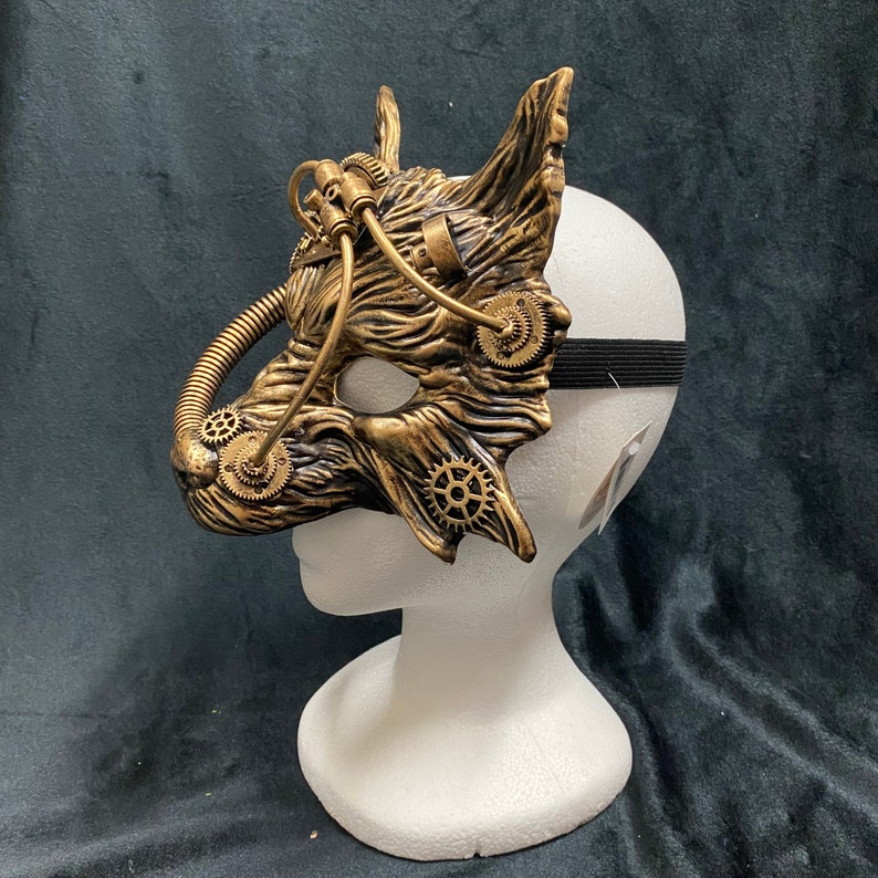 May include: A gold and black steampunk-style mask with gears and pipes. The mask is on a white mannequin head.