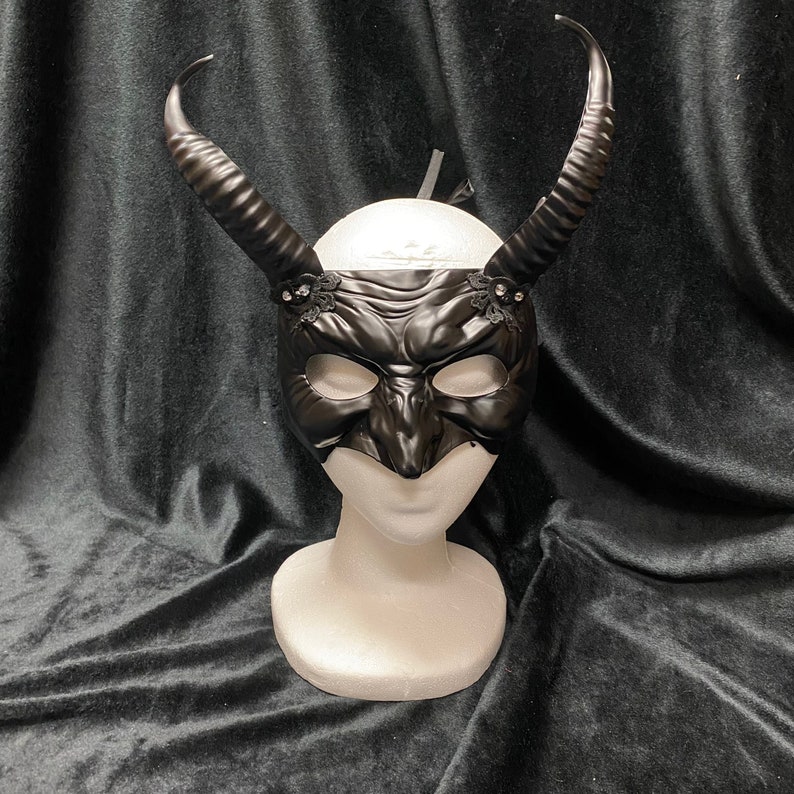 May include: A black masquerade mask with horns and lace accents. The mask has a detailed, textured design and is displayed on a white mannequin head.