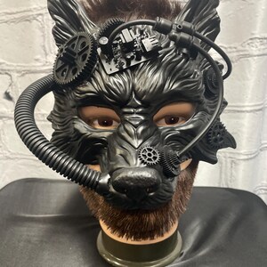 May include: A black wolf mask with steampunk details, including gears, pipes, and a circuit board. The mask is on a mannequin head with brown hair.
