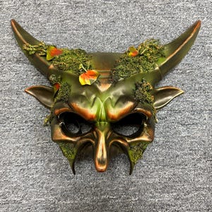 May include: A detailed mask with a horned design, featuring a blend of green, bronze, and gold tones. The mask has a textured appearance with added moss and leaf accents. The horns curve upwards, and the eye openings are prominent.
